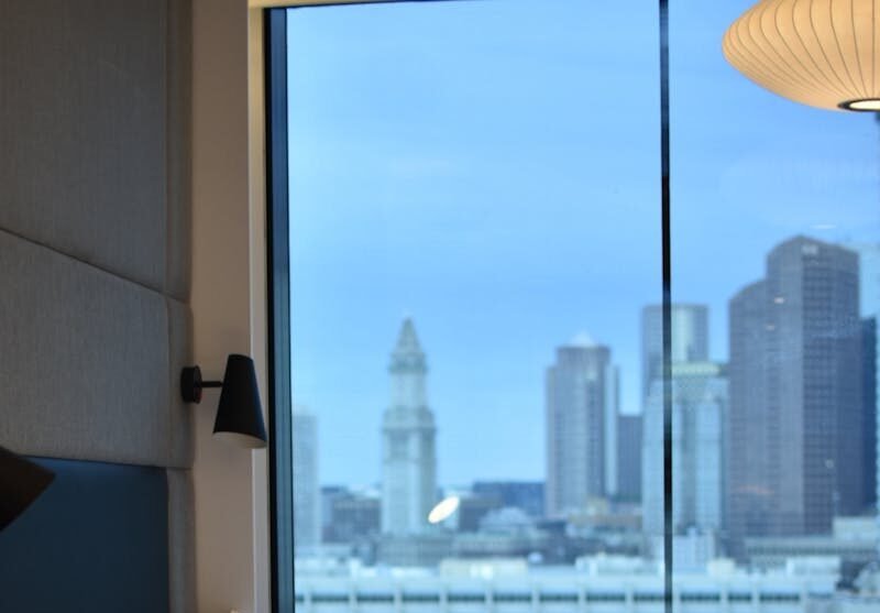 Modern hotel room overlooking Boston skyline, featuring luxury bedding.